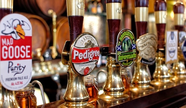 Close-up of beer taps at a bar with brass fittings and colorful circular brand logos like Pedigree and Mad Goose in view, on a wooden counter.